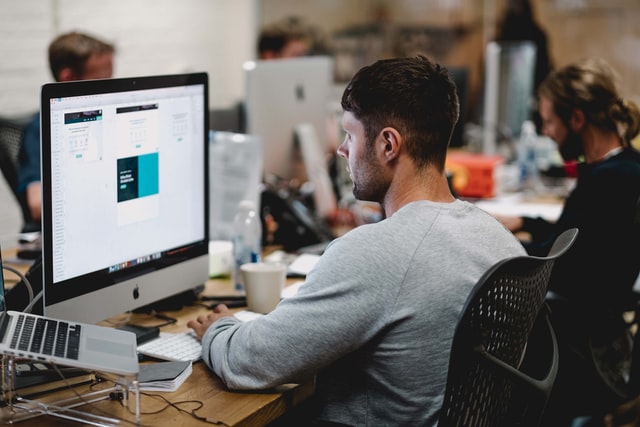 A man in a grey sweatshirt sitting on a chair in front of iMac.